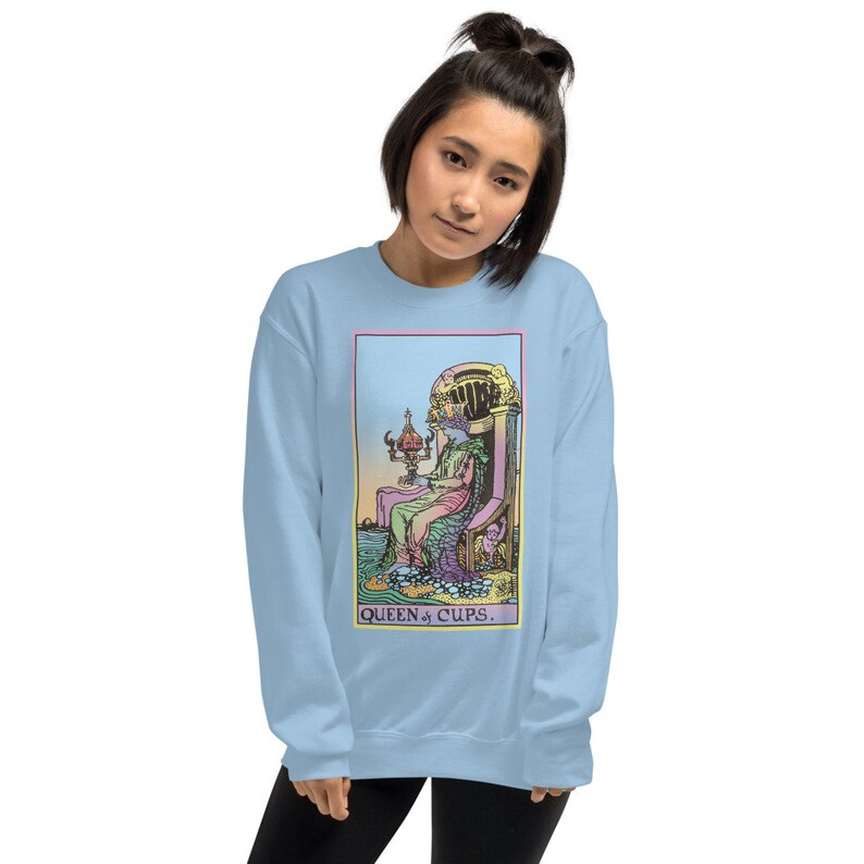 Puede incluir: Una sudadera de color azul claro con una ilustraci&oacute;n colorida de la carta del tarot de la Reina de Copas. La carta muestra a una mujer sentada en un trono con una copa en la mano. El texto "QUEEN of CUPS." est&aacute; impreso debajo de la imagen.