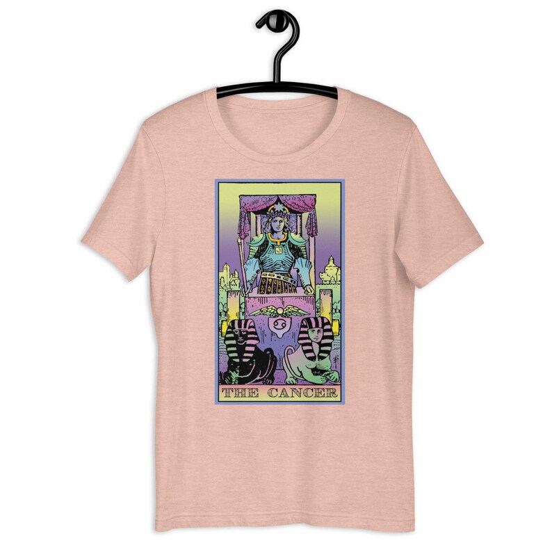 Puede incluir: Una camiseta rosa con un dise&ntilde;o de carta de tarot colorido que presenta el signo del zod&iacute;aco C&aacute;ncer. La carta muestra una figura sentada en un trono con dos figuras de esfinge en la parte inferior.