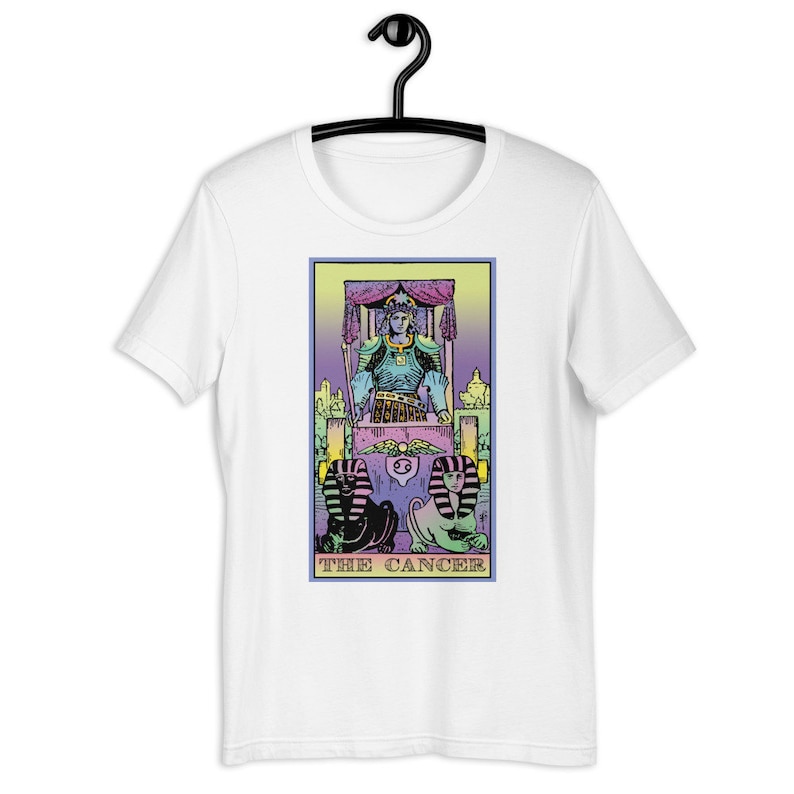 Puede incluir: Camiseta blanca con una ilustraci&oacute;n colorida de una carta de tarot, El C&aacute;ncer. La carta presenta una figura en un trono con dos figuras arrodilladas en la parte inferior.