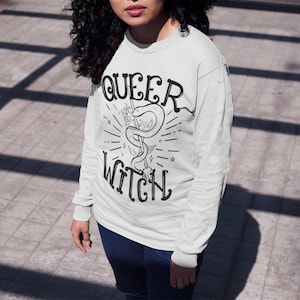 May include: White long-sleeve shirt with the text "QUEER WITCH" in black lettering. The shirt features a black and white illustration of a snake wrapped around a staff with flowers.