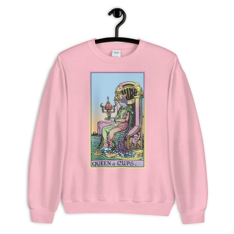 Puede incluir: Sudadera rosa con una ilustraci&oacute;n colorida de la carta del tarot de la Reina de Copas. La carta representa a una mujer sentada en un trono con una copa en la mano. El texto "QUEEN of CUPS." est&aacute; impreso debajo de la imagen.