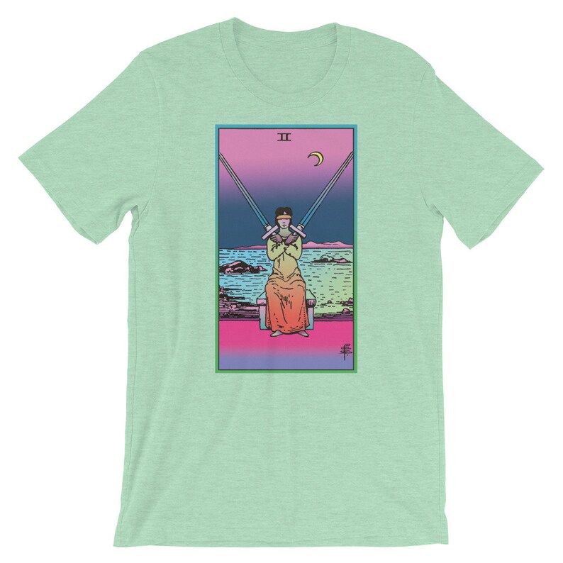 Op de afbeelding: Een groen T-shirt met een tarotkaartontwerp met een geblinddoekte figuur die twee zwaarden vasthoudt. De kaart is getiteld "II" en heeft een halve maan in de rechterbovenhoek.