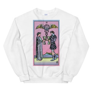 May include: White sweatshirt with a colorful illustration of the Two of Cups tarot card. The card features two figures holding cups, a winged lion head, and the Roman numeral II.