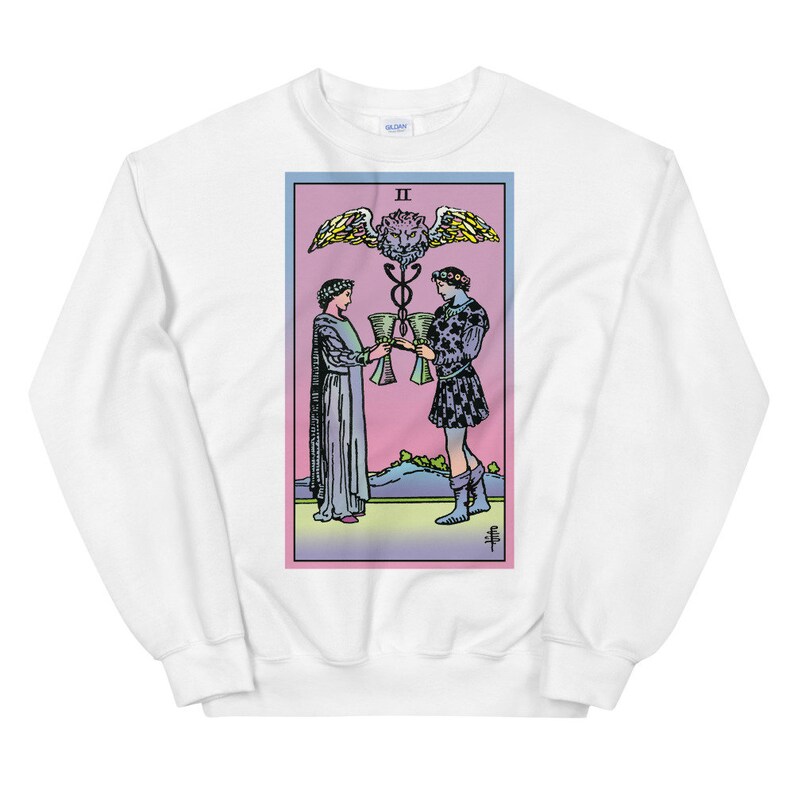 May include: White sweatshirt with a colorful illustration of the Two of Cups tarot card. The card features two figures holding cups, a winged lion head, and the Roman numeral II.