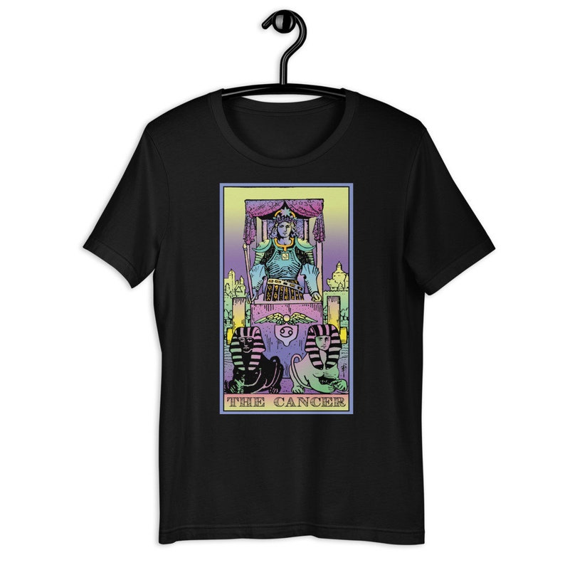 Puede incluir: Camiseta negra con una ilustraci&oacute;n colorida de la carta del tarot El C&aacute;ncer. La carta presenta una figura con una corona sentada en un trono con dos figuras de esfinge en la parte inferior. El texto "THE CANCER" est&aacute; impreso debajo de la imagen.