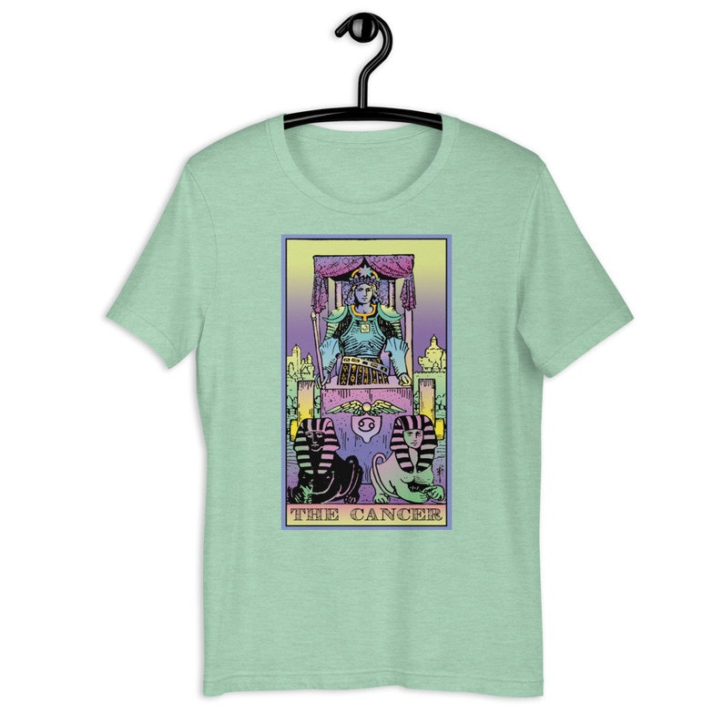 Puede incluir: Una camiseta verde con una ilustraci&oacute;n en colores pastel de una carta de tarot que representa El C&aacute;ncer. La carta muestra una figura sentada en un trono con dos figuras parecidas a esfinges en la parte inferior. El texto "THE CANCER" est&aacute; impreso debajo de la imagen.
