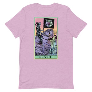 Puede incluir: Camiseta rosa claro con un dise&ntilde;o de carta del tarot. El gr&aacute;fico representa una figura esquel&eacute;tica a caballo, sosteniendo una bandera con una rosa. La palabra "DEATH" est&aacute; impresa debajo. El dise&ntilde;o est&aacute; enmarcado en un borde verde claro.