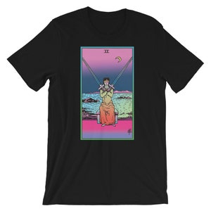Op de afbeelding: Zwart T-shirt met een kleurrijke illustratie van de tarotkaart Twee van Zwaarden. De kaart toont een figuur met twee zwaarden die hun ogen bedekken, zittend op een troon met een halve maan op de achtergrond.