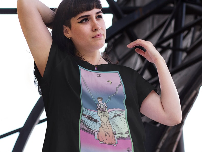 Op de afbeelding: Zwart T-shirt met een kleurrijk tarotkaartontwerp met een persoon in een gewaad die op een rotsachtige kust staat. De kaart is gemarkeerd met "II" en heeft een halve maan in de rechterbovenhoek.