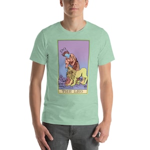 May include: A light green t-shirt with a graphic of a tarot card depicting The Leo. The card features a woman with a halo and a lion. The card is surrounded by a purple, pink, and yellow border. The text "THE LEO" is printed at the bottom of the card.