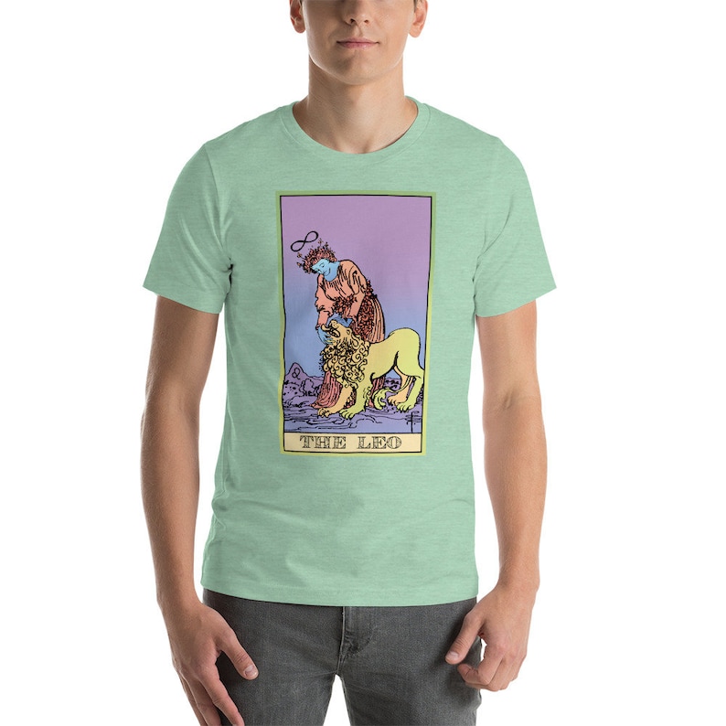 May include: A light green t-shirt with a graphic of a tarot card depicting The Leo. The card features a woman with a halo and a lion. The card is surrounded by a purple, pink, and yellow border. The text "THE LEO" is printed at the bottom of the card.