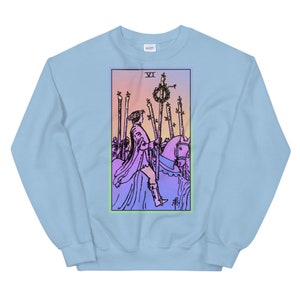 May include: Light blue sweatshirt with a graphic print of the Six of Wands tarot card. The card features a rider on horseback with a staff in hand, surrounded by other staffs. The background is a gradient of pink, orange, and yellow.