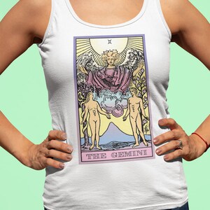 May include: White tank top with a colorful illustration of The Gemini tarot card. The card features a winged angel with two figures standing on either side.