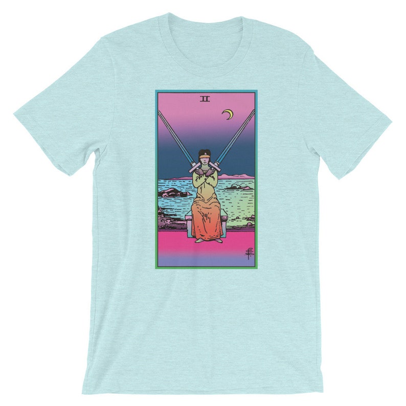 Op de afbeelding: Een lichtblauw t-shirt met een kleurrijke illustratie van de tarotkaart Twee van Zwaarden. De kaart toont een figuur met een blinddoek die twee zwaarden gekruist over zijn borst vasthoudt. De achtergrond is een verloop van roze, paars en blauw met een halve maan aan de hemel.