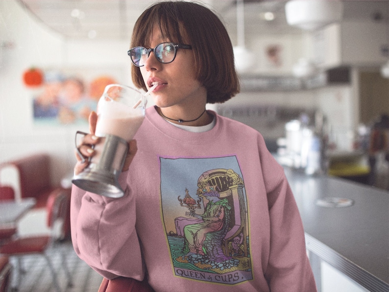 Puede incluir: Sudadera rosa con un estampado gr&aacute;fico de la carta del tarot de la Reina de Copas. La carta muestra a una mujer sentada en un trono con una copa en la mano. El texto "QUEEN of CUPS" est&aacute; impreso debajo de la carta.