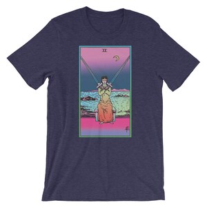 Op de afbeelding: Een marineblauw t-shirt met een kleurrijke illustratie van de tarotkaart Twee van Zwaarden. De kaart toont een figuur met een blinddoek die twee zwaarden gekruist over zijn borst vasthoudt.