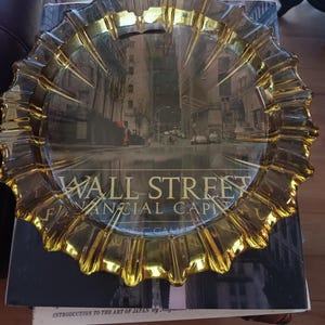 May include: A clear glass ashtray with a gold-colored, scalloped edge. The center of the ashtray features a photograph of Wall Street with the text "WALL STREET FINANCIAL CAPITAL". The ashtray is resting on a book.