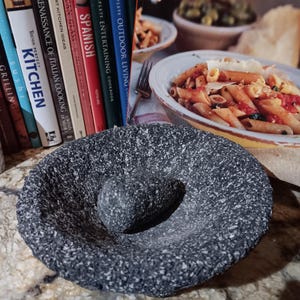 May include: A dark gray mortar and pestle set. The mortar is a bowl-shaped vessel with a rough, textured surface. The pestle is a rounded, cylindrical stone. The background includes cookbooks and a plate of pasta.