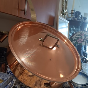 May include: A copper-colored pot with a matching lid. The pot has a brass handle and a brass hanging loop. The lid has a brass handle. The pot is sitting on a countertop with blue and white china.