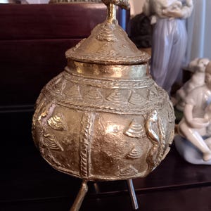 May include: A gold metal decorative stand with a round top and three legs. The stand is decorated with intricate carvings and a bird finial.