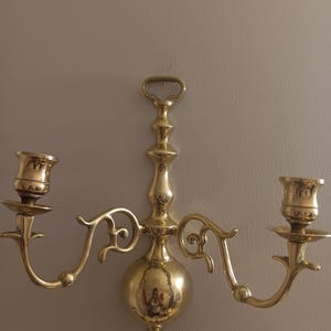 May include: A brass wall sconce with two candle holders. The sconce has a central decorative element with a handle at the top and curved arms extending to the candle cups. The finish is shiny and reflective.