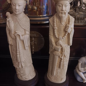 May include: Two ivory-colored statues of Asian figures. The statue on the left depicts a woman in ornate robes, while the one on the right portrays a man with a beard and hat. Both are mounted on dark wooden bases, showcasing detailed carvings.