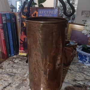 May include: A tall, cylindrical copper bucket with a black, curved handle. The bucket has a rustic, aged appearance with visible texture and a slightly tarnished finish. The handle is attached to the top rim, suggesting it's designed for carrying or hanging. The bucket is likely for decorative or functional use.