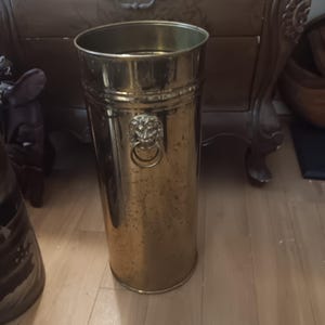 May include: A tall, cylindrical brass umbrella stand with a polished metallic finish. The exterior features a decorative lion head motif on the side, which incorporates a circular ring handle. The surface shows subtle signs of natural patina.