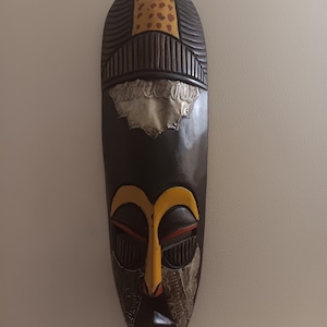 May include: A long, dark wooden mask with a pointed bottom. The mask features a yellow and orange face with silver accents. The top of the mask has a yellow and brown patterned design. The mask is likely for decorative purposes.