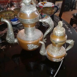 May include: Two ornate silver and copper teapots with intricate designs. The larger teapot has a silver lid and a chain attached to the handle. The smaller teapot has a silver lid and a silver spout.