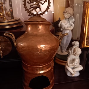 May include: A copper pot with a decorative brass lid featuring a dancing figure with multiple arms. The pot has a round body and a base with a hole in the center.