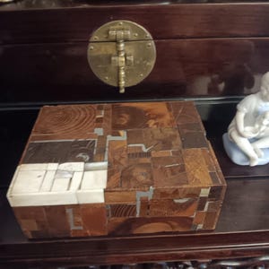 May include: A square wooden box with a mosaic design. The box is made of various wood pieces in shades of brown, tan, and white. The box is sitting on a dark brown wooden surface. The box has a hinged lid and a brass clasp.