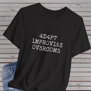 Op de afbeelding: Een zwart t-shirt met korte mouwen en witte tekst op de borst met de tekst 4D4P7 1MPROV153 0V3RCOM3. Het t-shirt heeft een ronde hals en wordt getoond naast een deel van een blauwe spijkerstof met een zichtbare metalen knoop en zak.