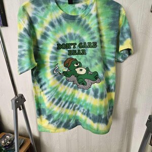 May include: A tie-dye t-shirt in green, yellow, and blue. The shirt has a graphic of a green bear smoking a cigarette, lying on a cloud, with the text "DON'T CARE BEAR" above it.