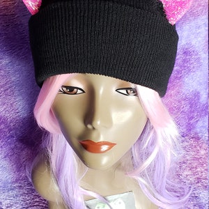 May include: A black knit beanie with pink glitter cat ears. The hat is displayed on a mannequin head with pink and purple hair. The background is a textured purple fabric.