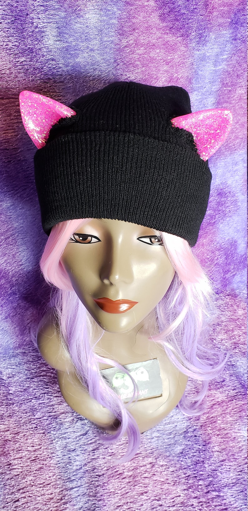 May include: A black knit beanie with pink glitter cat ears. The hat is displayed on a mannequin head with pink and purple hair. The background is a textured purple fabric.