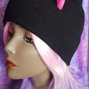 May include: Black knit beanie with pink glitter cat ears. The hat is worn by a mannequin with pink and purple wig. The background is a purple and pink fabric.