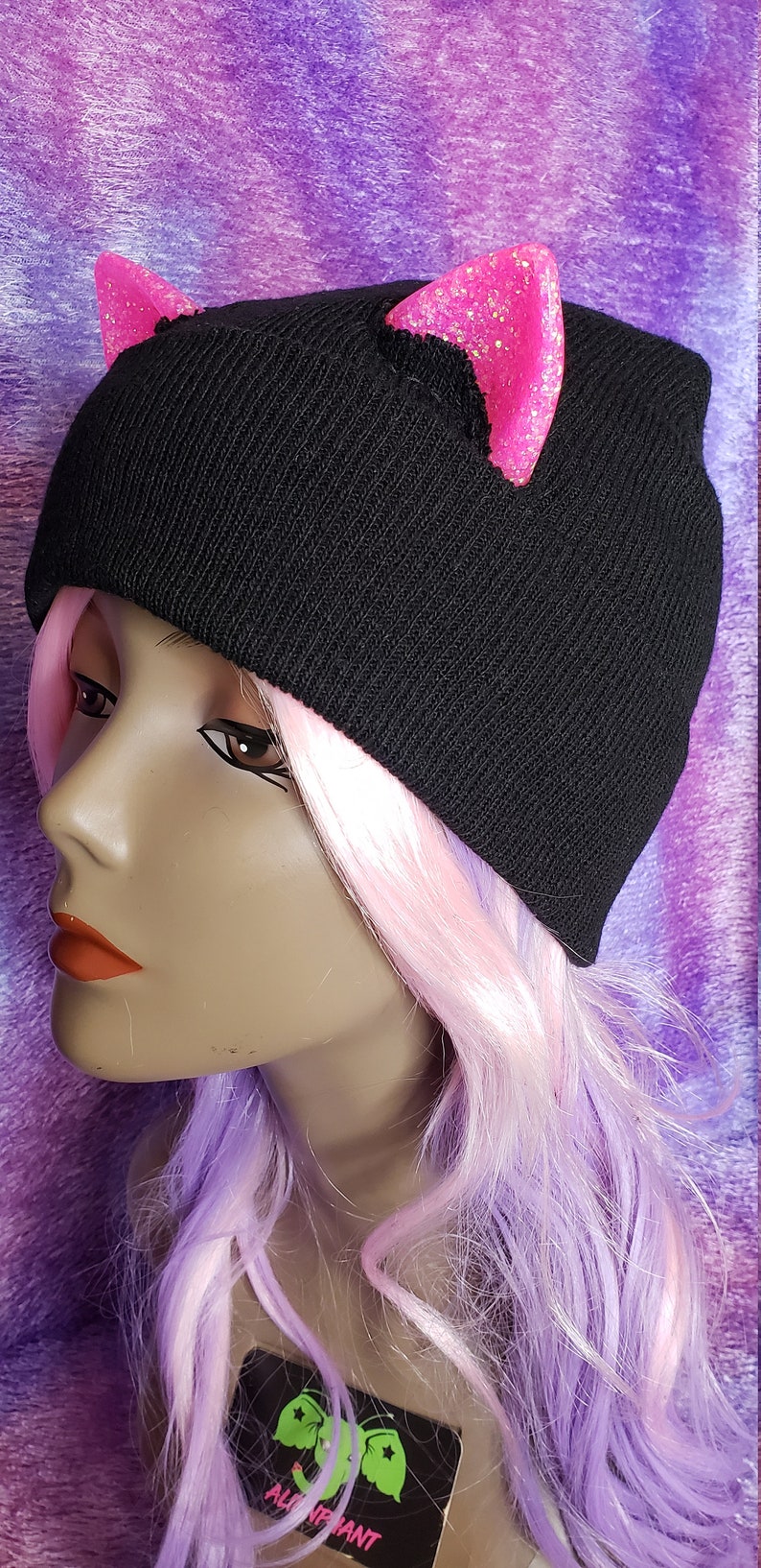 May include: Black knit beanie with pink glitter cat ears. The hat is worn by a mannequin with pink and purple wig. The background is a purple and pink fabric.