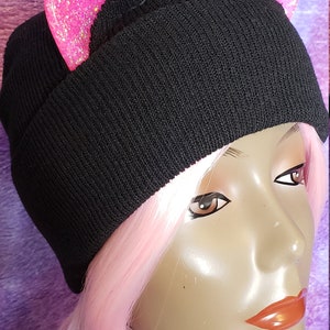 May include: Black knit beanie with pink glitter cat ears. The hat is worn by a mannequin with pink hair and a purple background. The cat ears are a bright pink color and add a playful touch to the hat.