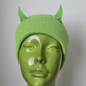 May include: A lime green knit beanie featuring two glossy, translucent lime green devil horns attached to the top. The fabric has a ribbed texture and a slightly distressed, frayed appearance around the base of each horn, creating a distinct look.
