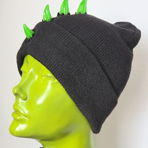 May include: A black knit beanie featuring four bright green, glossy, cone-shaped spikes protruding from the top. The hat has a folded cuff and a textured fabric construction, showcasing a row of spikes along the crown of the headwear.