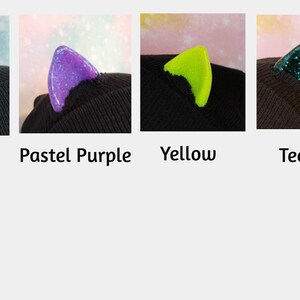 May include: Four close-up images of cat ears in red, pastel purple, yellow, and teal. Each ear is attached to a black beanie. The red and teal ears have glitter. The text below each image indicates the color.