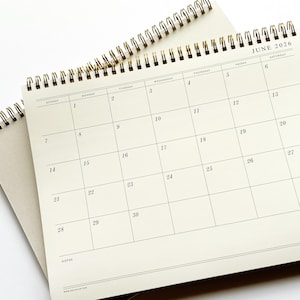 May include: A close-up of a spiral-bound calendar open to June 2026. The calendar pages are a light beige color with black grid lines and dates. The top right corner displays the month and year. The spiral binding is gold-colored.