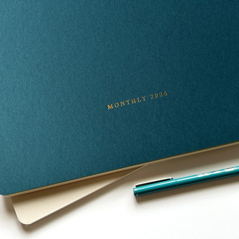 May include: A teal-colored planner with the words "MONTHLY 2026" in gold lettering. A teal pen rests on the corner of the planner. The planner is open, revealing a cream-colored page.