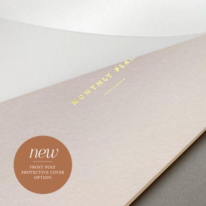 May include: A close-up of a light pink planner with the words "MONTHLY PLAN" in gold lettering. A brown circle with the word "new" and "FROST POLY PROTECTIVE COVER OPTION" is in the lower left corner. The planner is on a white surface.