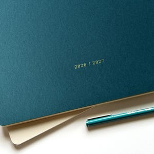 Pode incluir: Uma agenda verde-azulado escura com capa texturizada apresentando o texto 2026 / 2027 gravado em dourado. A agenda está sobre uma página cor de creme e uma caneta verde-azulado a condizer com clipe prateado está posicionada na diagonal.