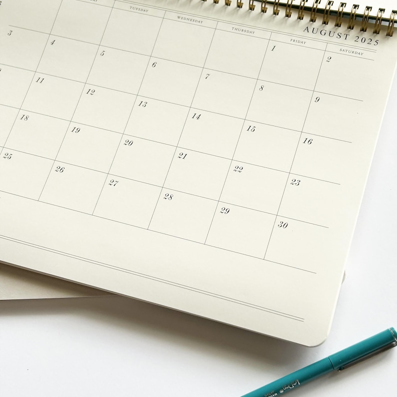May include: A close-up of an open spiral-bound calendar for August 2025. The calendar pages are cream-colored with a grid layout. A teal pen rests at the bottom. The calendar is on a white surface.