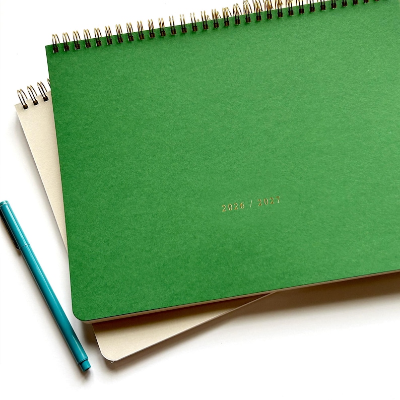 May include: A green spiral-bound notebook with gold lettering reading "2026 / 2027" and a teal pen. The notebook is on top of a beige notebook. The spiral binding is gold.