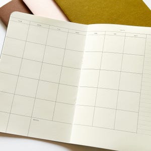 May include: An open planner with cream-colored pages featuring a black grid layout for a monthly calendar. The headers include SUN, MON, TUE, WED, THR, FRI, SAT, and MONTH. A notes section is labeled at the bottom left. Three closed notebooks in brown, pink, and olive green are visible behind the open planner.