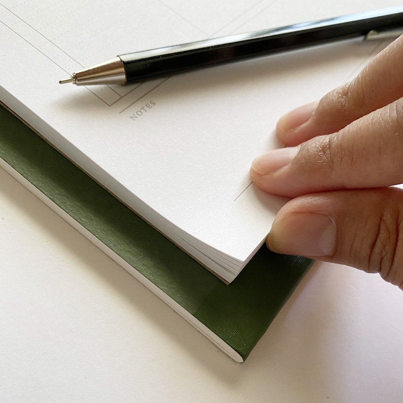 K&ouml;nnte beinhalten: Ein gr&uuml;nes Notizbuch mit einer umgeschlagenen wei&szlig;en Seite. Auf der Seite steht das Wort "NOTES". Ein schwarzer Stift liegt auf dem Notizbuch.